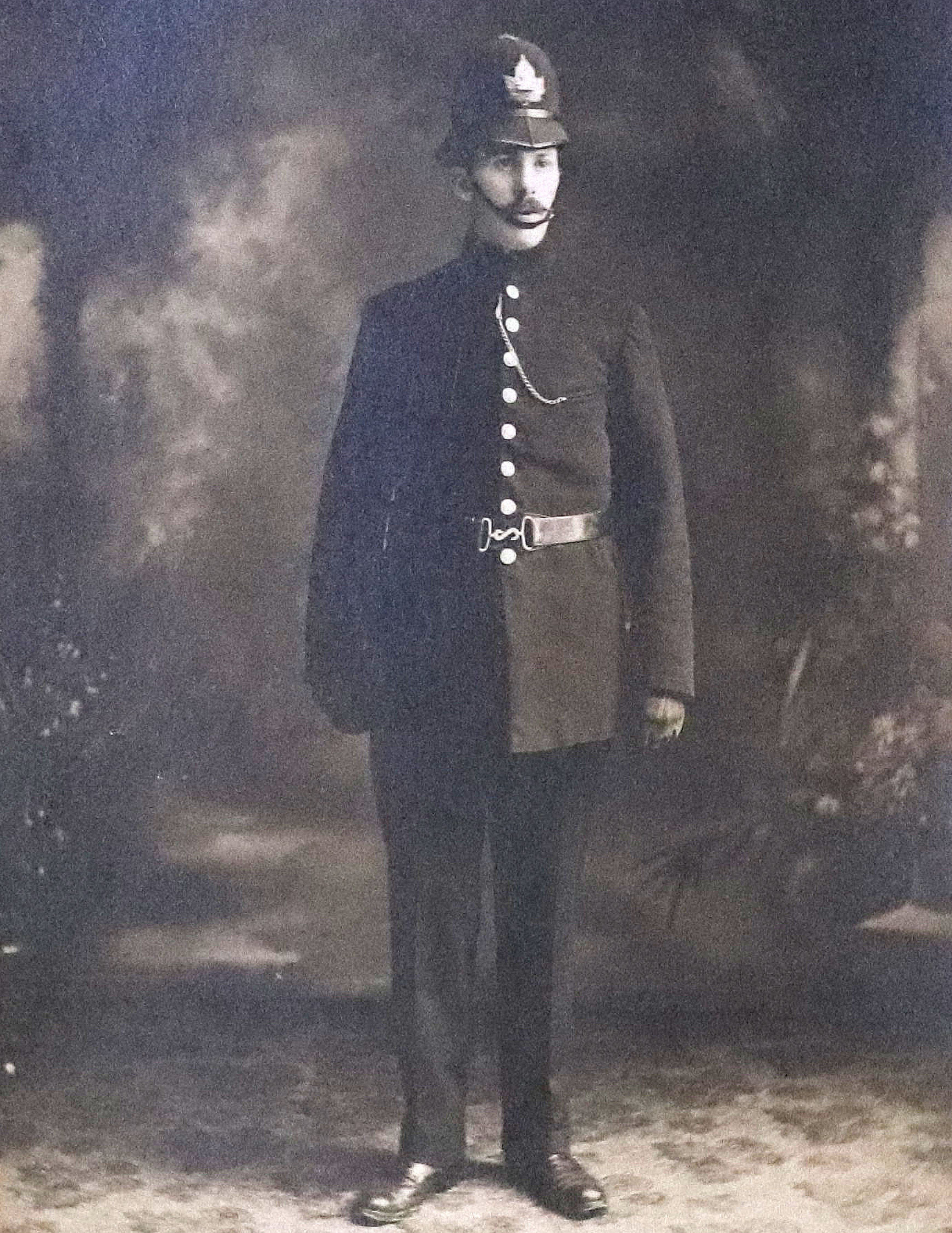 A man wearing a dark coloured police uniform. The coat features a row of brass buttons. He is wearing a rounded helmet with a chin strap. 