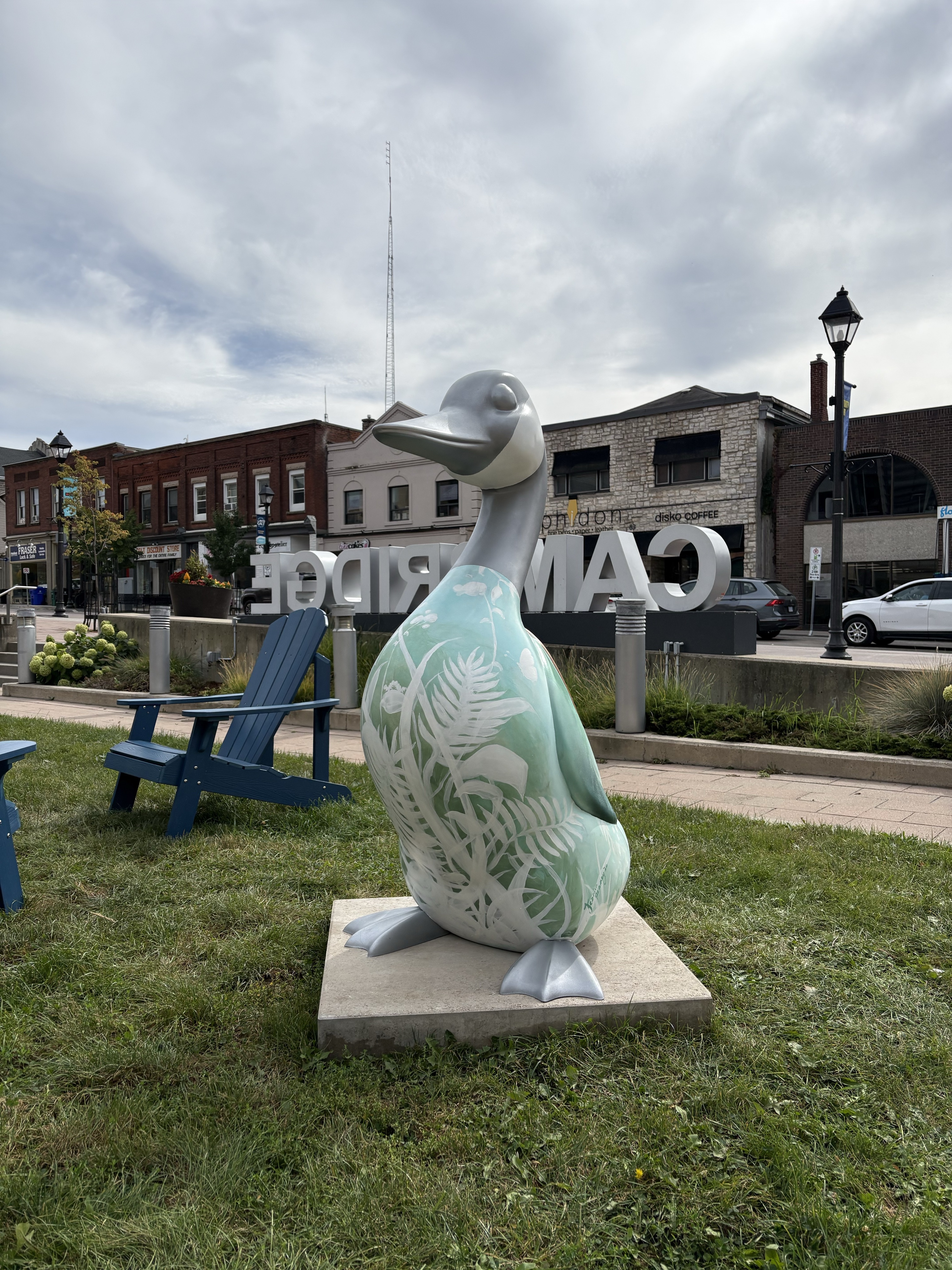 a 4.5-foot-tall fibreglass goose standing on a concrete base on green grass.  The goose's feet, neck, and head are painted silver and its torso light green with white silhouettes of plants