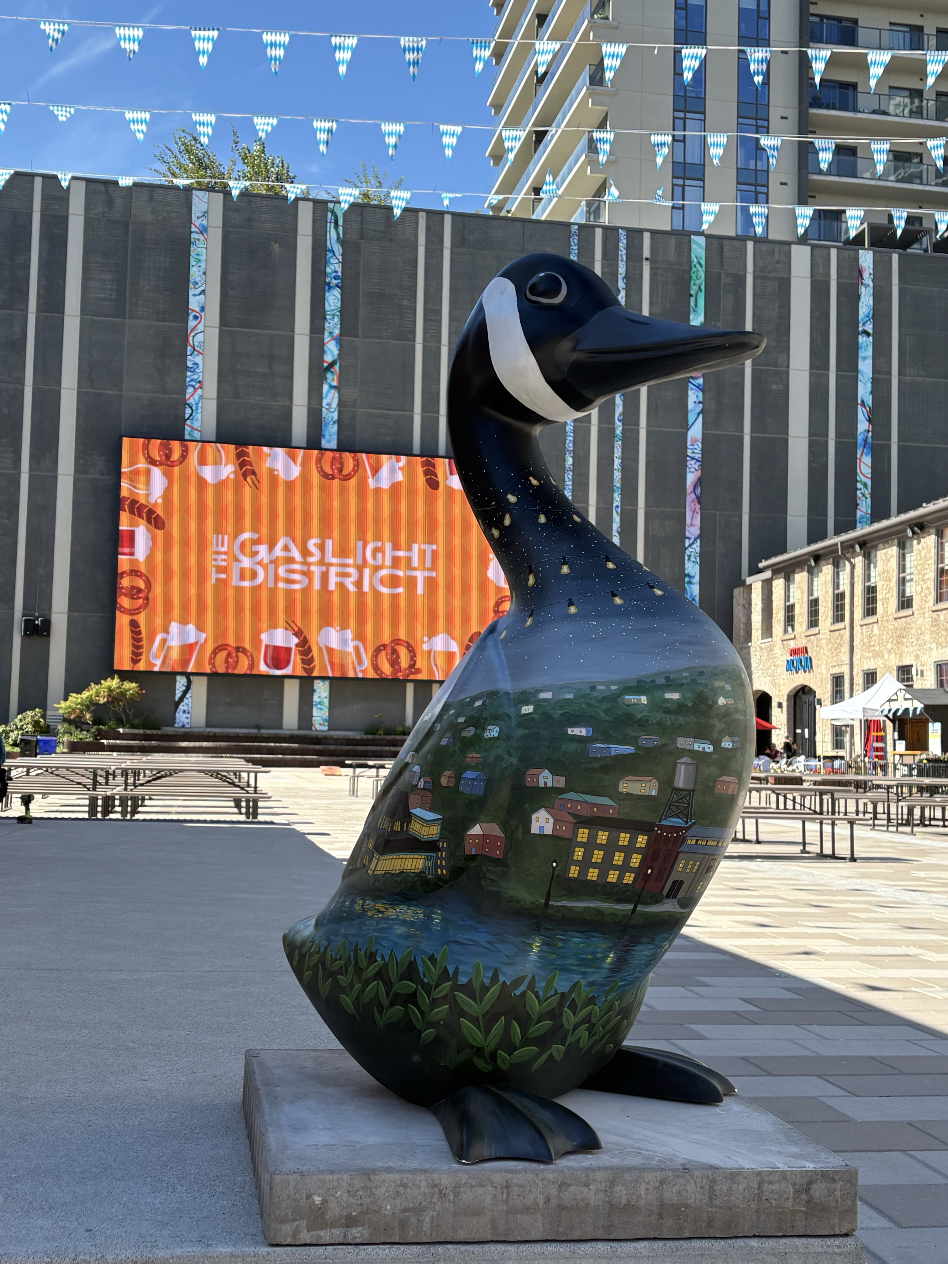 Large fibreglass goose sculpture painted with lanscape featuring buildings and a river below an evening sky. The goose is standing in front of large electronic billboard that reads 'the Gaslight District'.