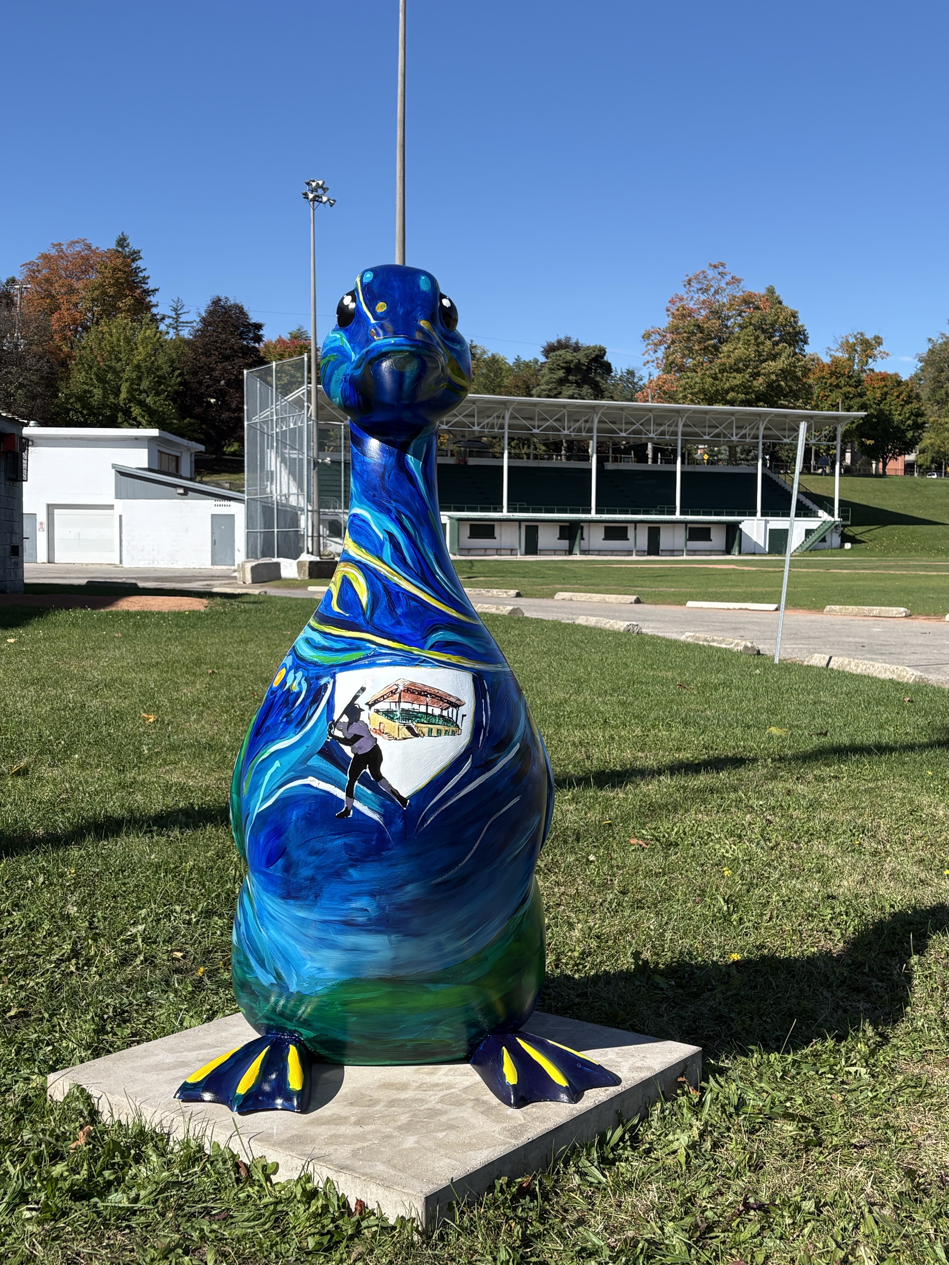 A 4.5-foot-tall fibreglass goose standing in front of the baseball field and grand stand. The goose is painted with organic marks in blue, green, turquoise and yellow with a crest on its chest displaying a baseball player and an image of the grand stand.