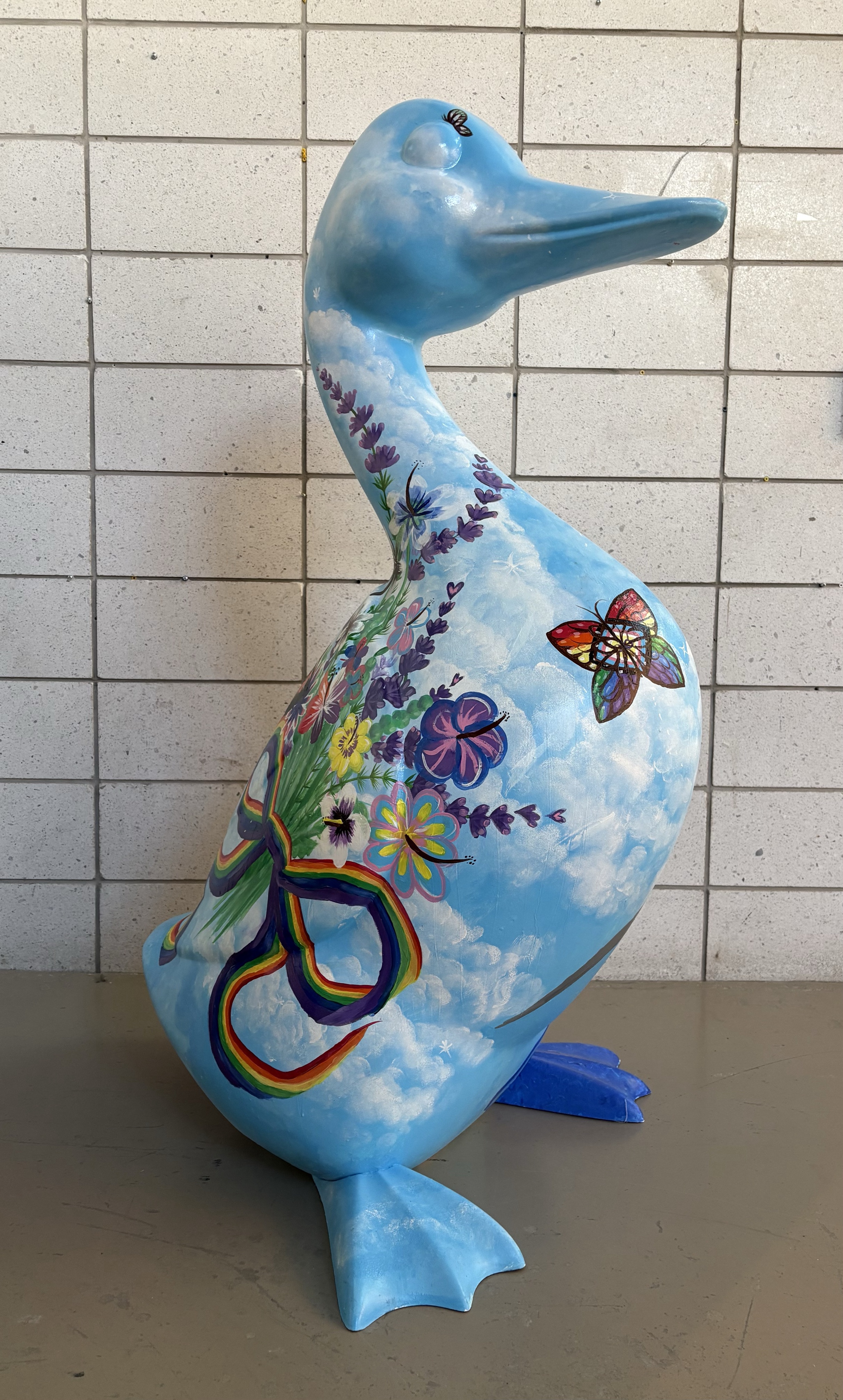 A 4.5-foot-tall fibreglass goose painted with a bouquet of colourful flowers tied with a rainbow ribbon, set in front of a cloudy blue sky.