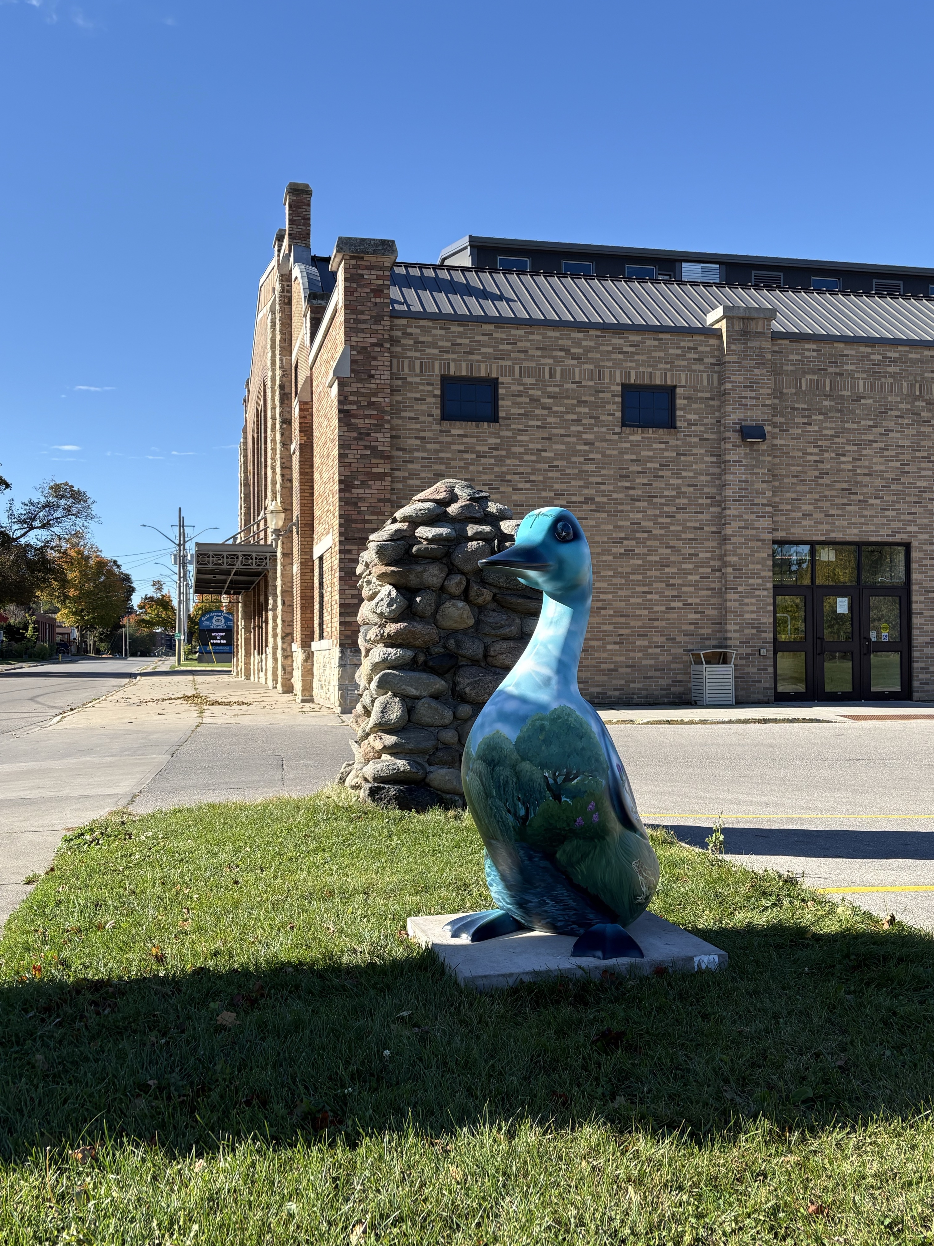 A 4.5-foot-tall fibreglass goose standing in front of a large yellow brick building.  The goose is painted with a landscape featuring a body of water below green willow trees and a cloudy blue sky.