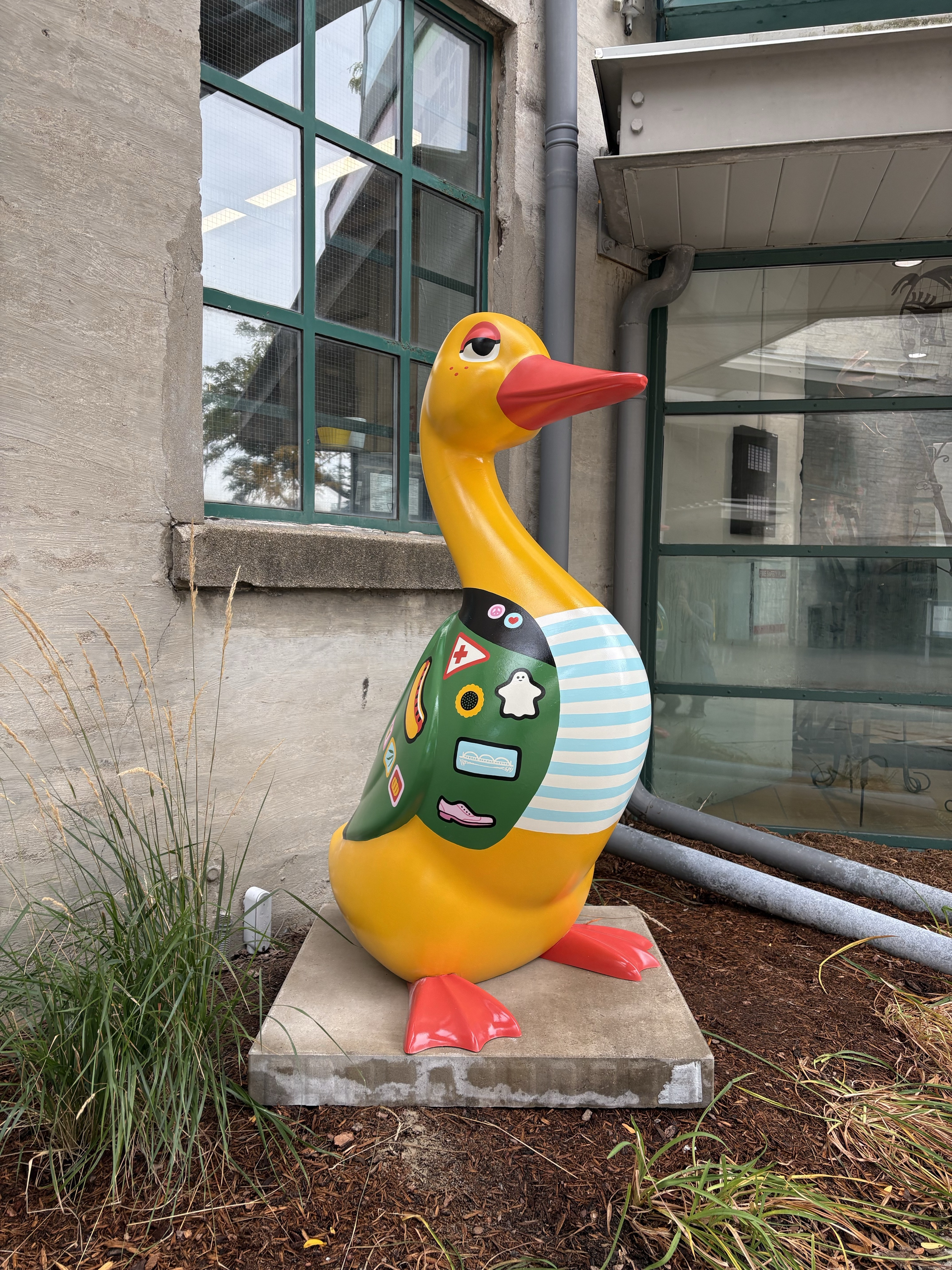 a 4.5-foot tall fibreglass duck painted golden yellow with orange beak and feet.  The duck is painted to look like it is wearing a light blue and white striped shirt under a green jacket with patches representing a ghost, a shoe, and a bridge