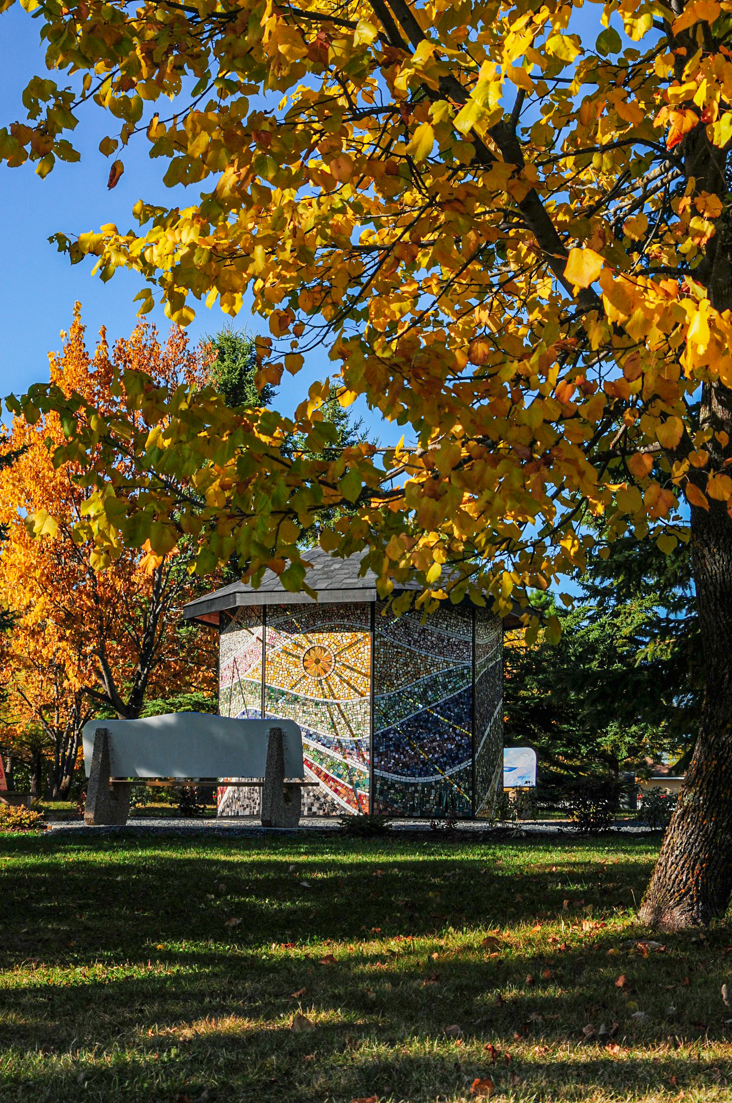 A colorful mosaic-covered structure in a park, surrounded by trees with vibrant autumn leaves in shades of yellow and orange, under a sunny sky.