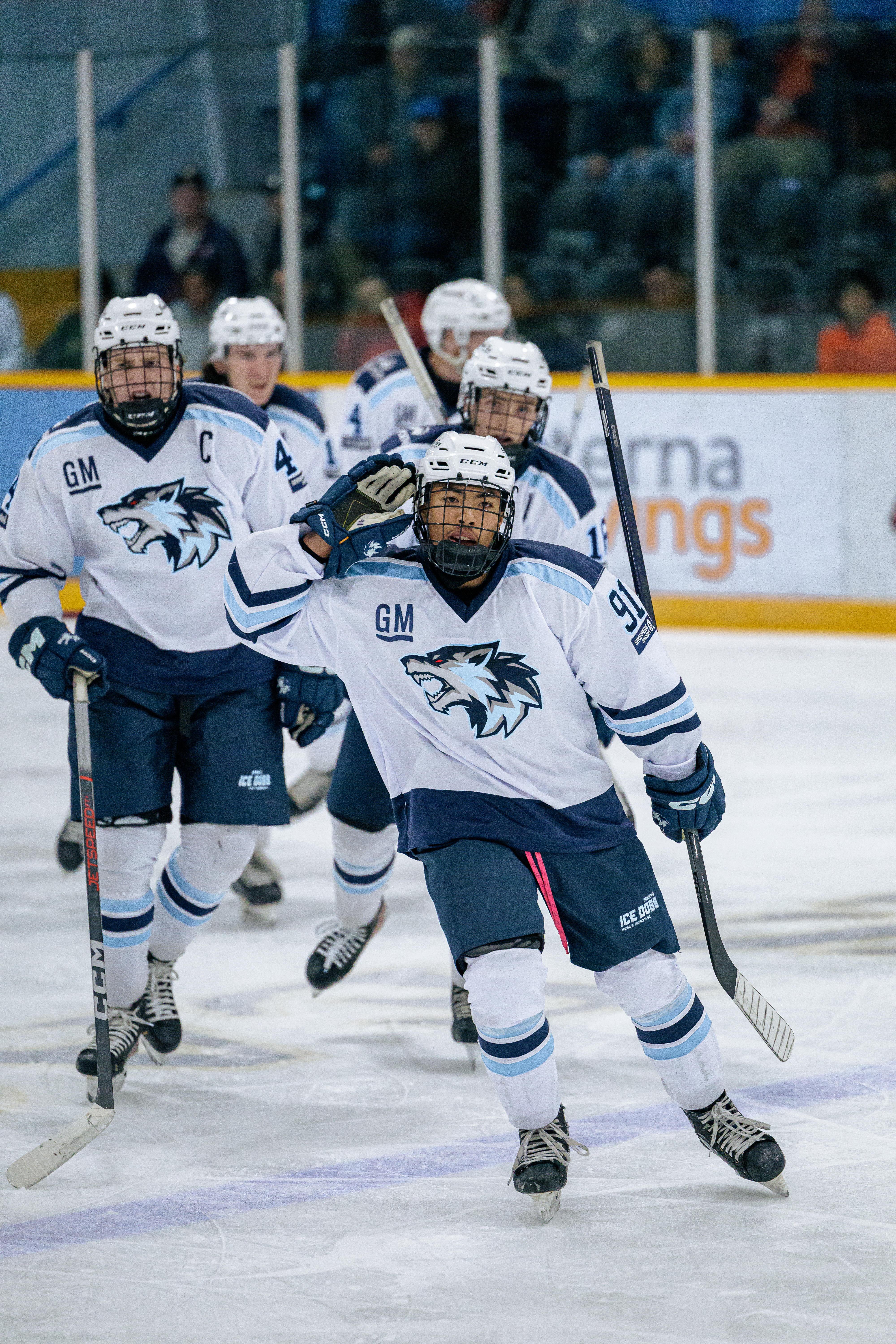 Hockey player in a white and blue dog-logo jersey celebrates on the ice while teammates follow behind during a game
