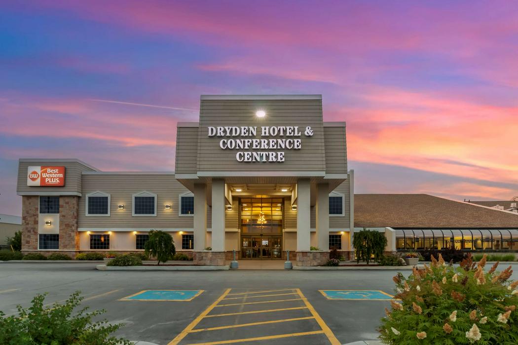 The Dryden Hotel & Conference Centre, a modern building with a "Best Western Plus" sign, illuminated entrance, and chandelier, set against a vibrant pink and purple sunset sky, with a parking lot and bushes in the foreground.