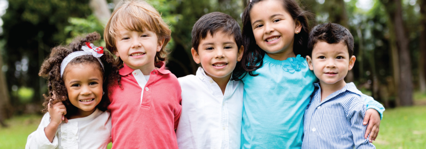 Five children are standing together and smiling in an outdoor setting with greenery