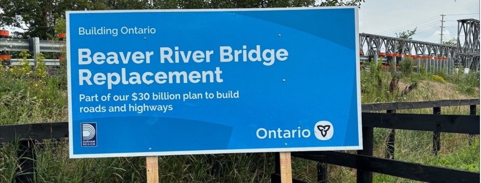A sign announcing the Beaver River Bridge Replacement, with the temporary Beaver River Bridge in the background.