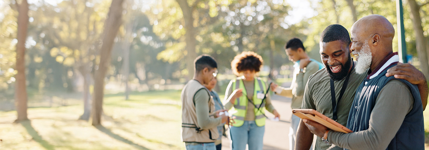A group of people in a park, with two individuals in the foreground looking at a tablet together. Other members are engaged in conversation and looking at clipboards. The setting is outdoors with trees and sunlight.