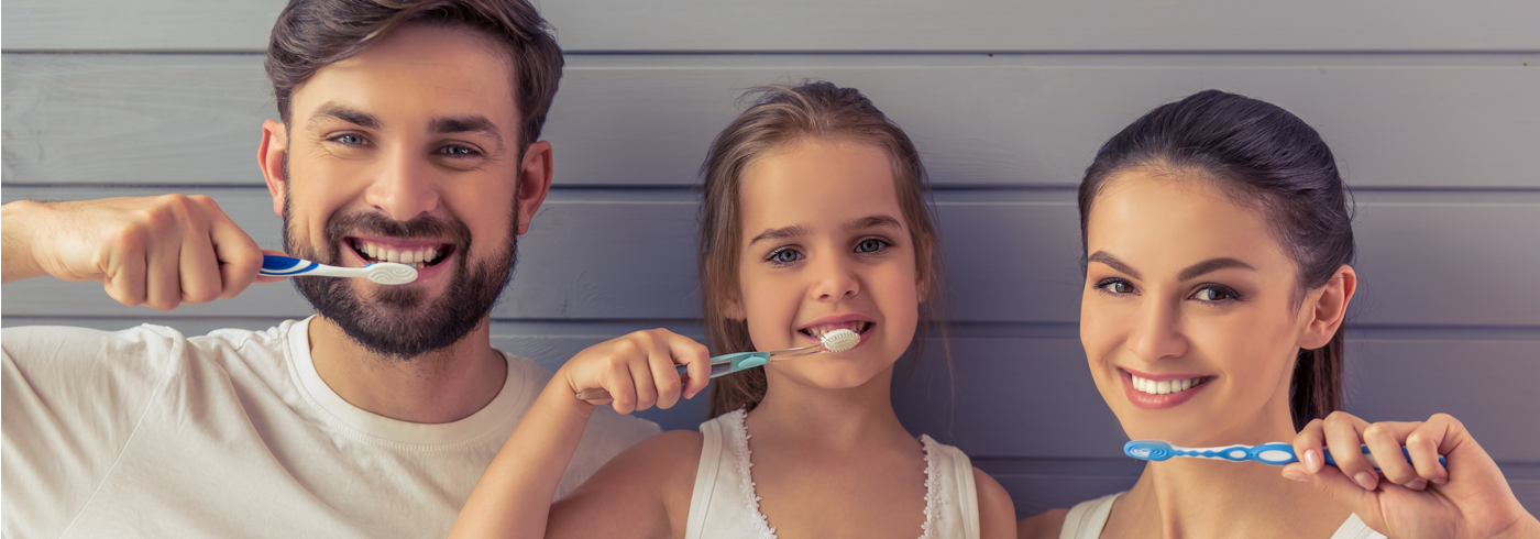 Three people smile while brushing their teeth with blue toothbrushes. They stand in front of a light wooden wall.