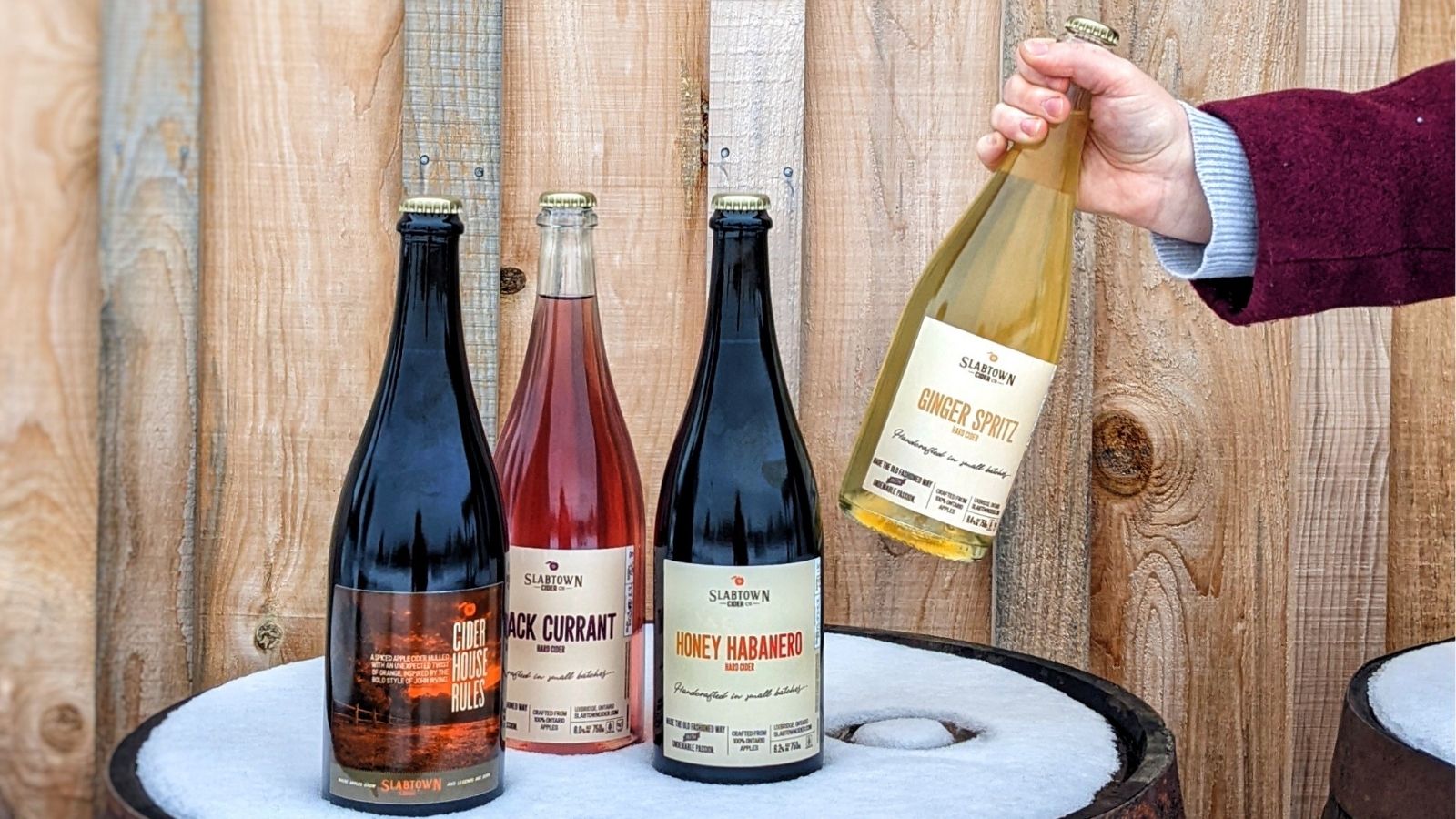 Image of bottles of cider sitting on top of a snowy barrel with one of the bottle being lifted up by an arm