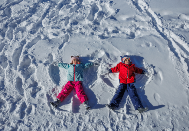 Two children wearing winter gear making snow angels next to each other.