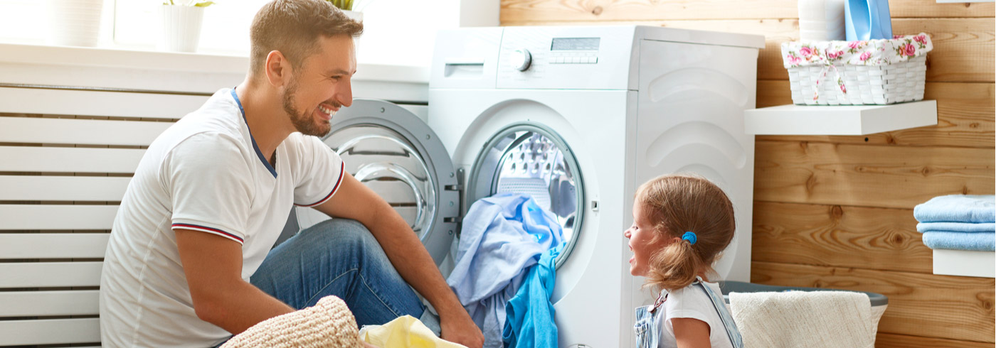 A person and a child are in a laundry room. The adult is crouching near an open washing machine filled with clothes. The child, smiling, is standing nearby holding a light blue cloth. The room has wood-paneled walls and shelves with towels and a basket.