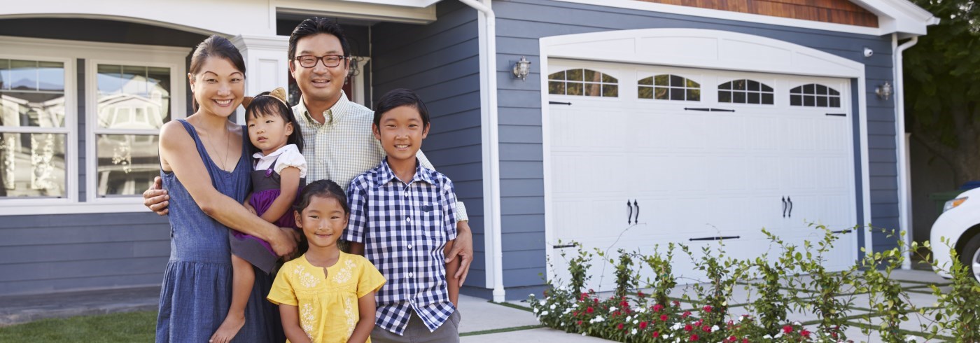 A group of people is smiling in front of a house with a blue exterior and a white garage door. Flowers line the walkway leading to the house.