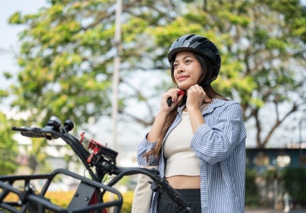 Person adjusts the chin strap of a helmet while standing beside an e-bike outdoors.