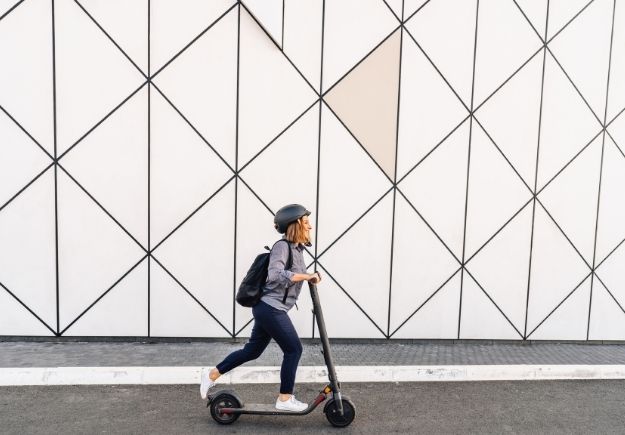 Person wearing a helmet and backpack riding an e-scooter along a sidewalk beside a modern geometric-patterned wall.