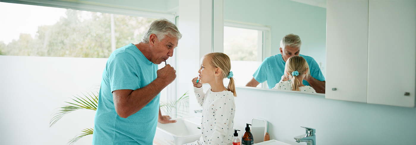 An older adult and a child are standing side by side in a bright bathroom, both brushing their teeth in front of a mirror.