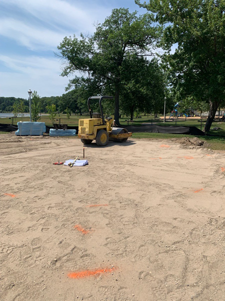 A construction site for a future splash pad shows a leveled sandy area with survey markings and a yellow roller compactor parked near the center. Trees, a playground, and a waterfront park are visible in the background under a partly cloudy sky.