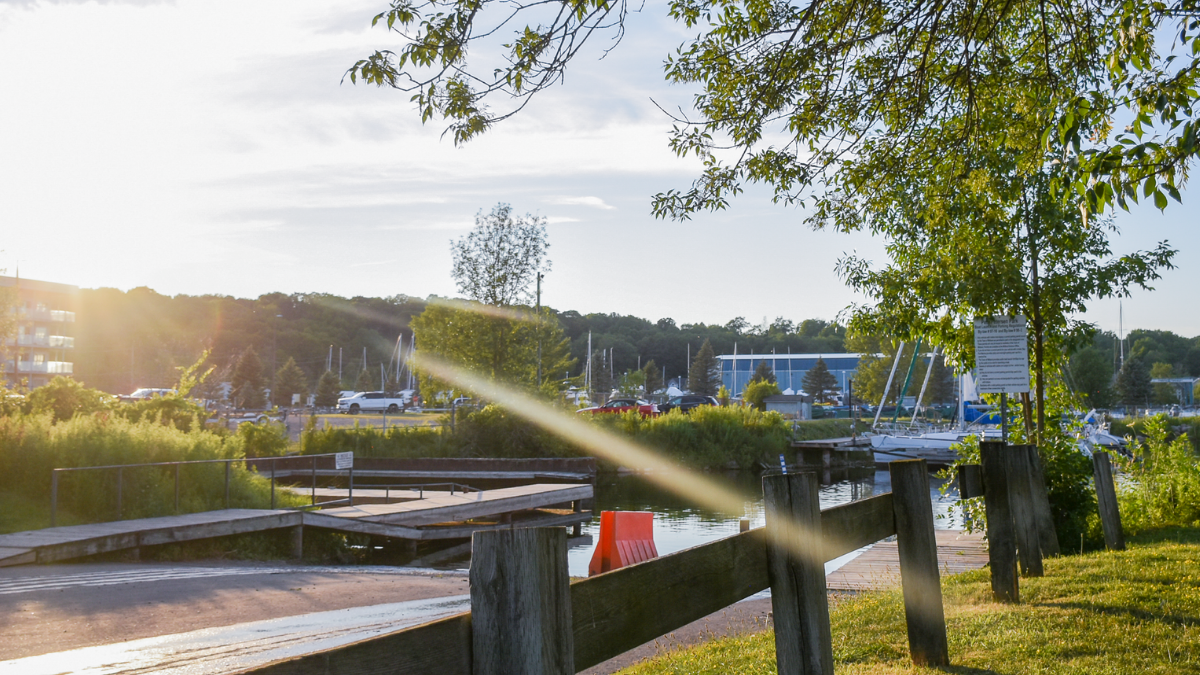 A sunlit waterfront boat launch with wooden docks, calm water, and marina boats in the background. Trees frame the scene, and a beam of sunlight streaks across the image during golden hour.