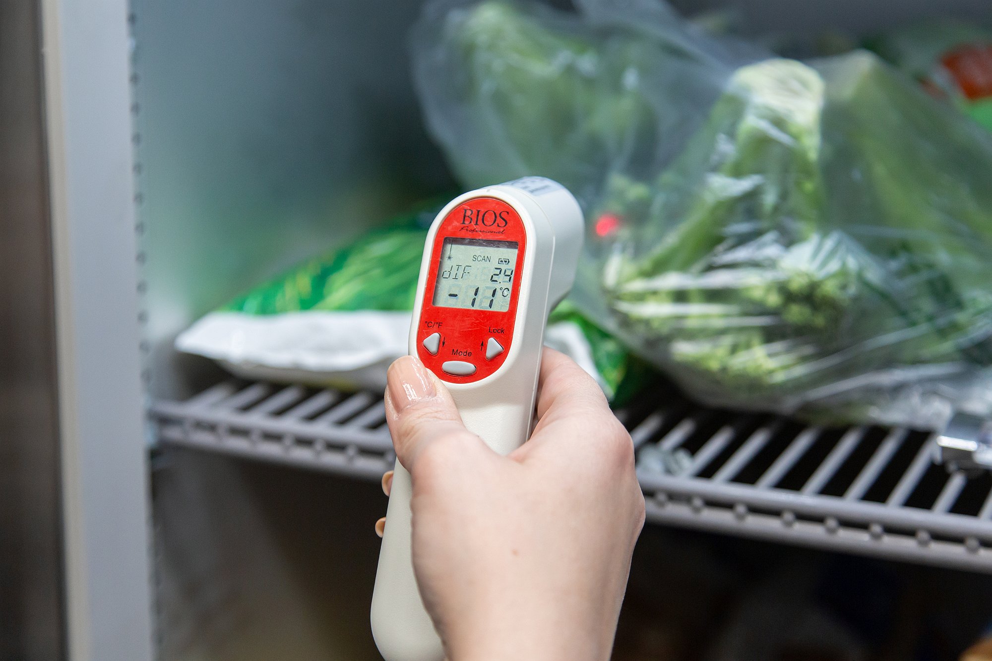A hand is holding a digital thermometer to check the temperature inside a fridge filled with produce