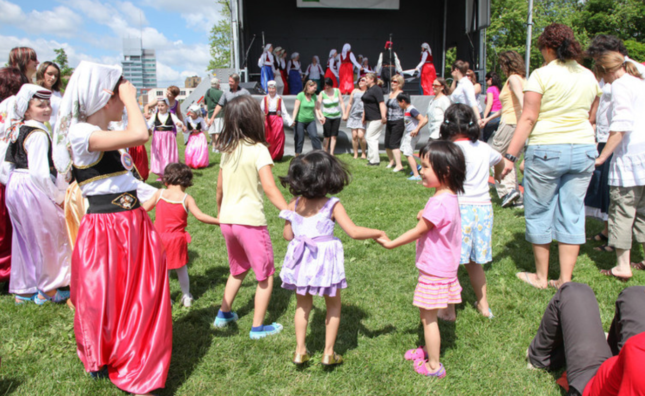 A circle of people of mixed ages and backgrounds dancing together while performers are on a stage in a background