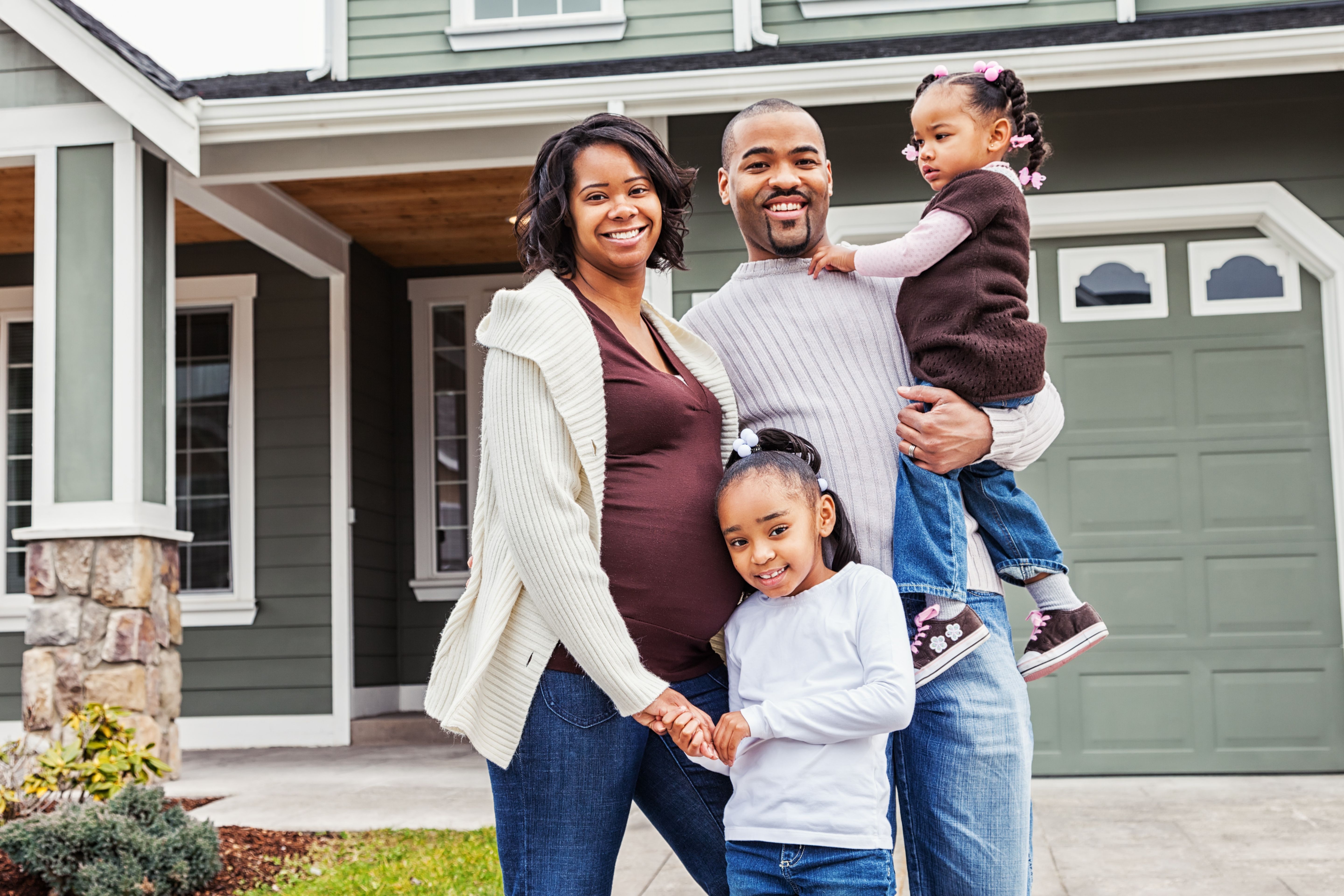 A young, black, family with a pregnant mother, a father, a school-aged daughter and a toddler standing in front of their house. 