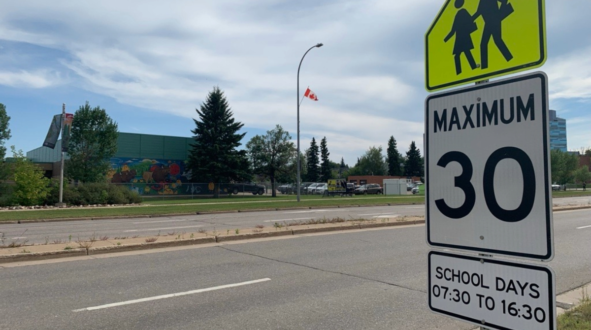 Image with white and yellow speed sign and a school in the background
