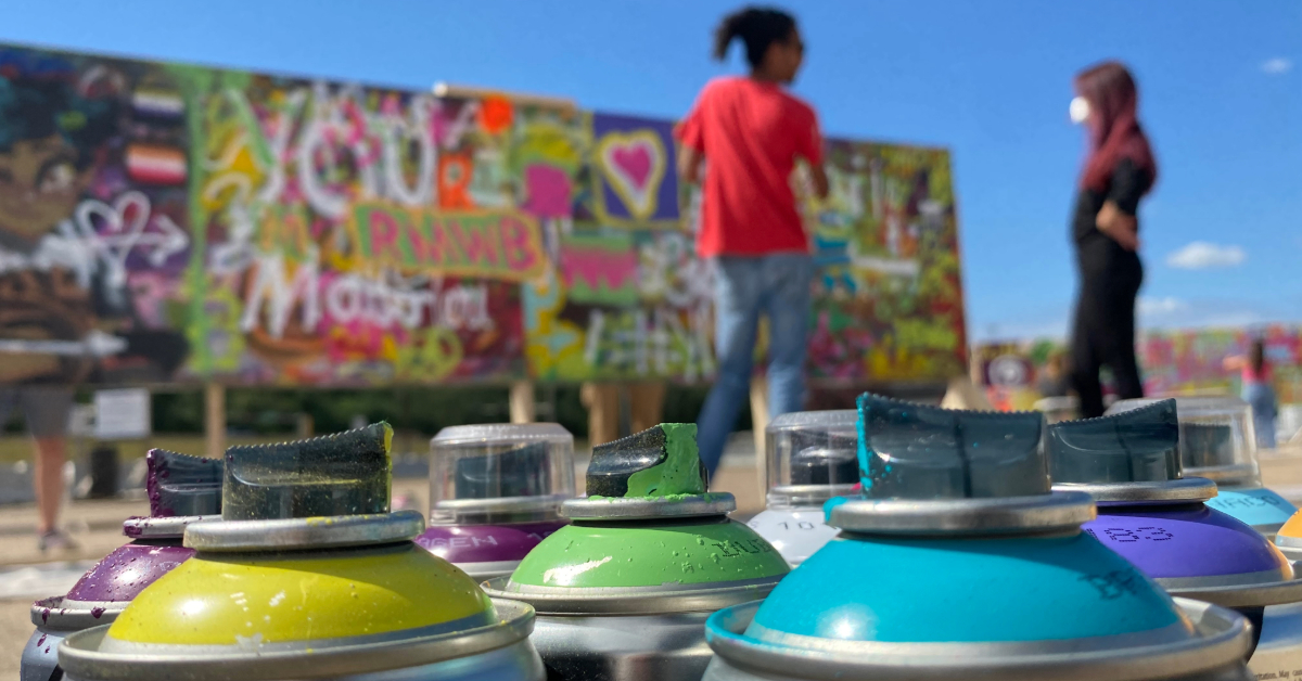 Spray paint cans of all different colours are in the forefront with artists in the background.