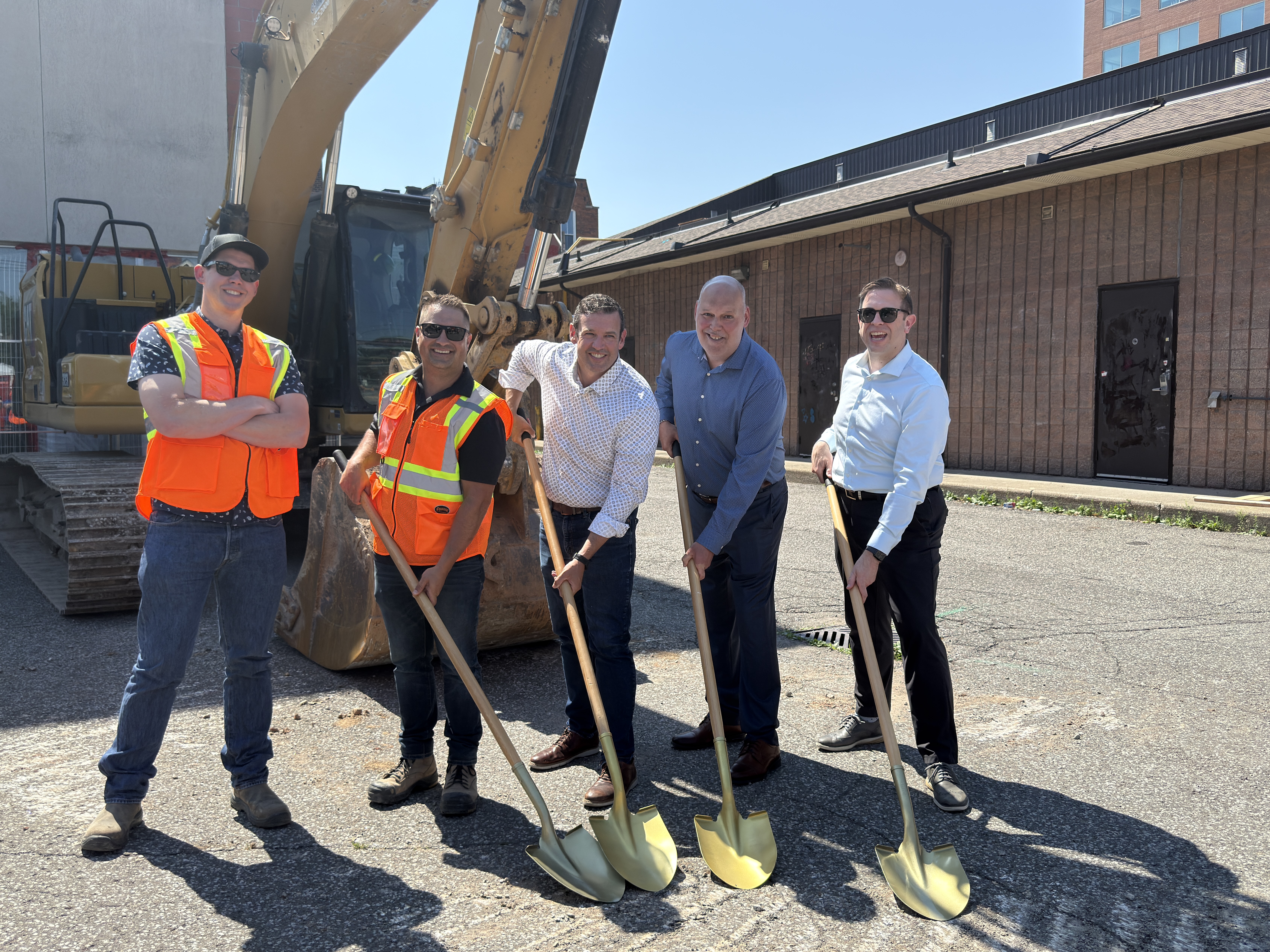 Five people stand on a construction site with golden shovels and a large excavator behind them, for a ceremonial groundbreaking.