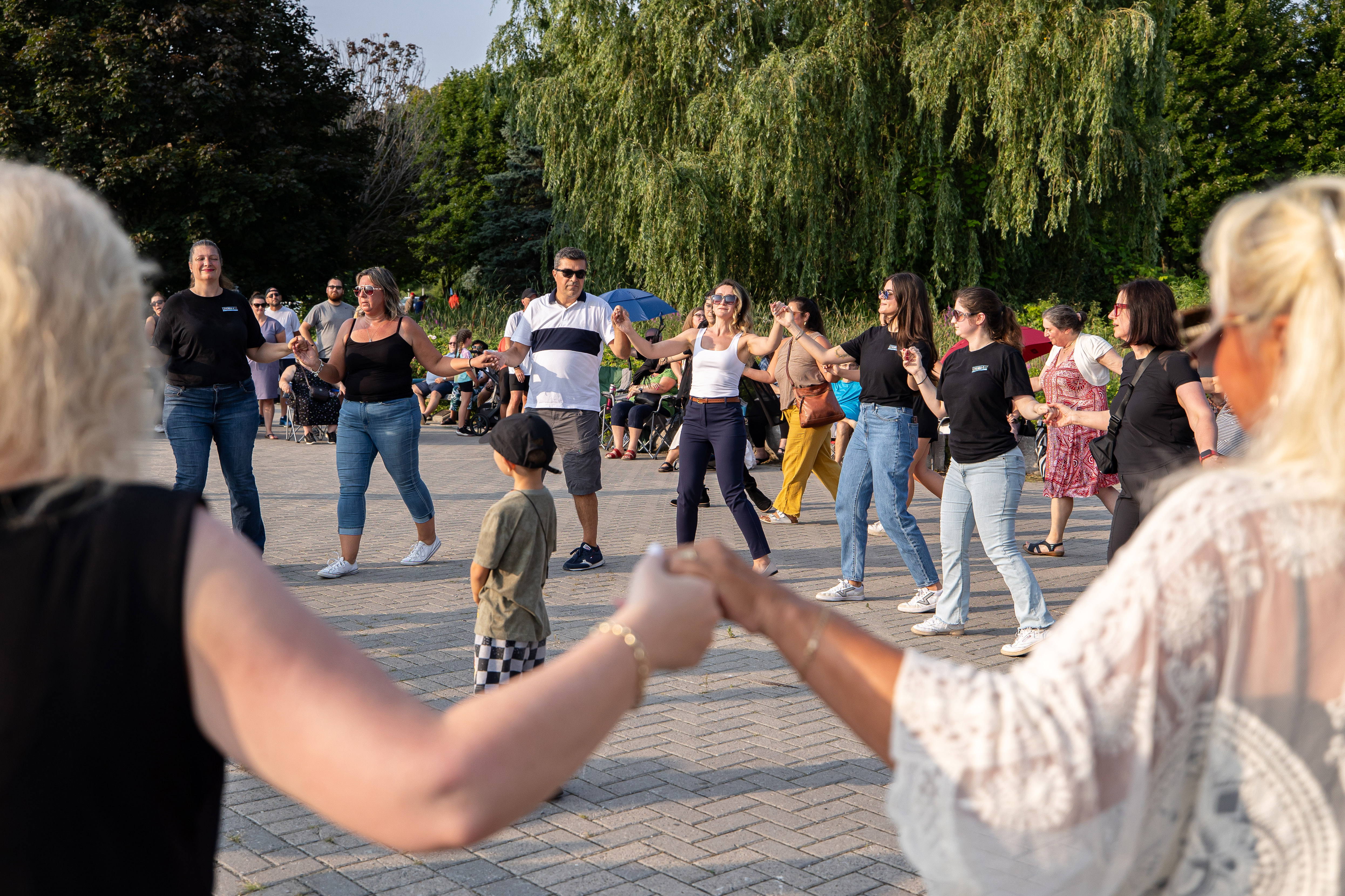 large group of people holding hands and dancing in traditional Greek dance with large trees in background 